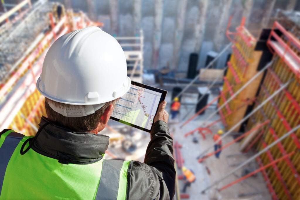 A construction worker reviewing project status on a tablet while overseeing a job site.