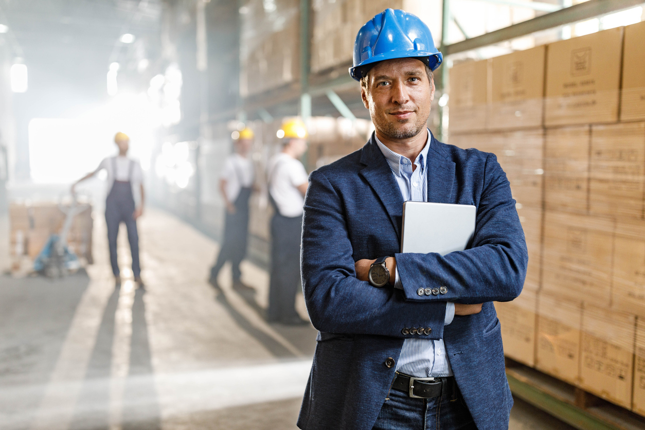 A man in business clothing wearing a hardhat and carrying a laptop stands in a warehouse with workers in overalls working behind him.